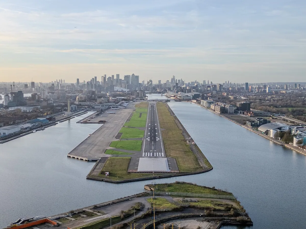 The photograph shows a wide aerial view of London City Airport, looking westwards along the single runway, with the River Thames and surrounding docklands clearly visible.